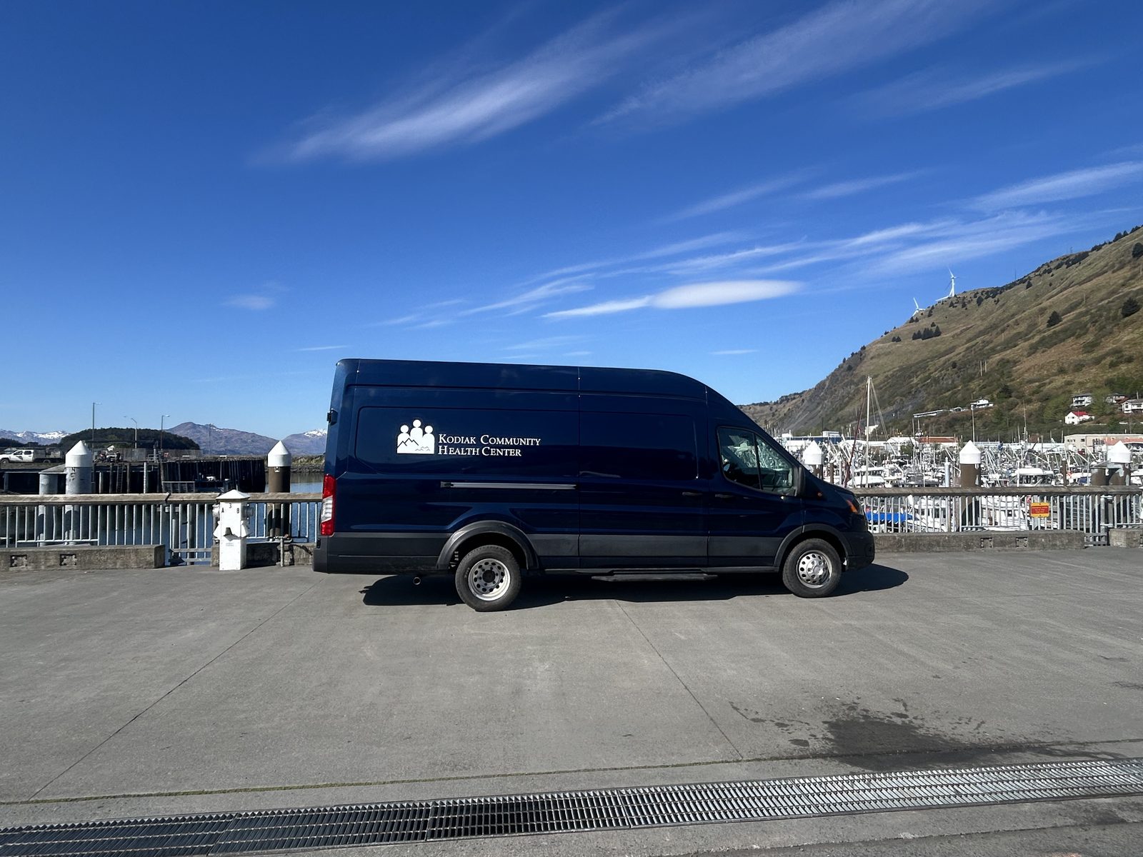 The mobile health unit parked at the Kodiak harbor, with fishing boats and hillside wind turbines in the background